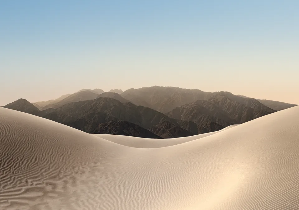 iPhone Duo default inner display Lock Screen wallpaper, light version: pale sand dunes and hazy mountains under a soft blue morning sky.